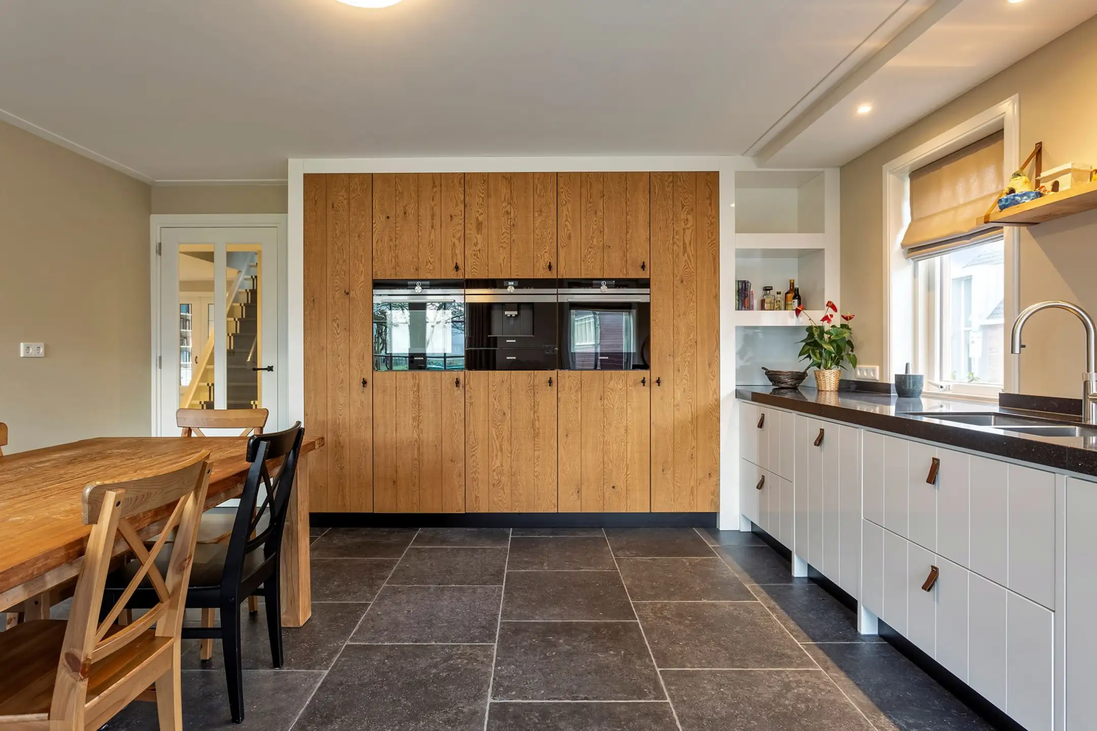 Vastgoedfotografie van een moderne landelijke keuken met een grote houten kastenwand, ingebouwde ovens, een donkere natuurstenen vloer en een eethoek.