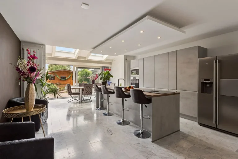 Vastgoedfotografie van een luxe, moderne woonkeuken met een kookeiland, barstoelen, inbouwspots, een marmeren vloer en een schuifpui naar de tuin.